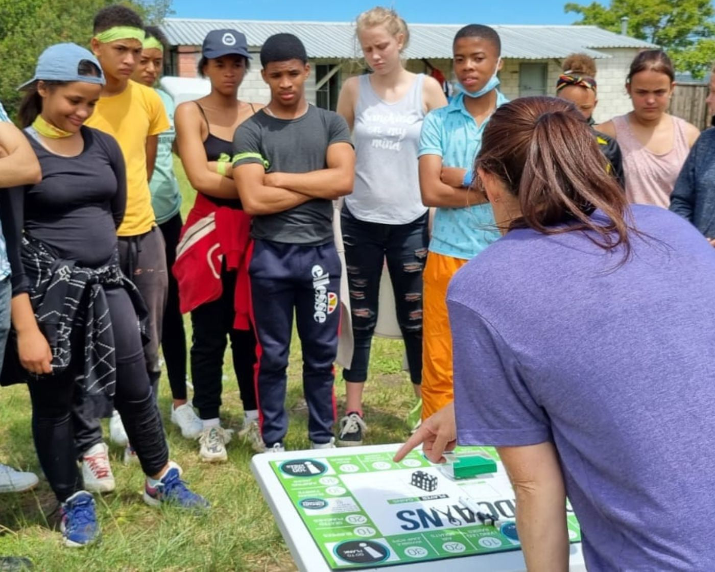 A group of young people standing outdoors on grass, gathered around a board game with a woman in a purple shirt pointing at the game board. The scene is set in a sunny outdoor environment with a house and trees in the background.