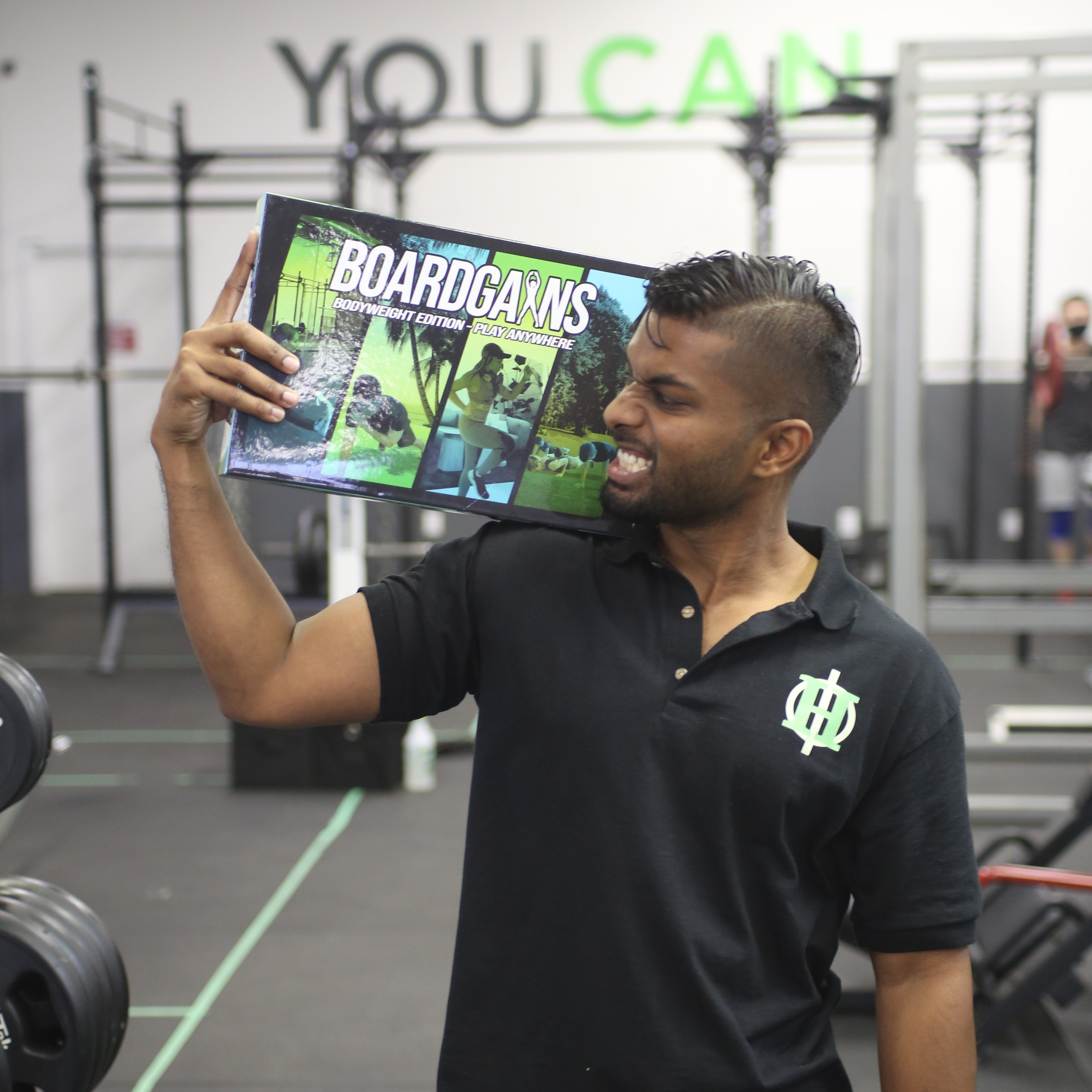 A man in a black polo shirt with a green logo holds up a board game box titled 'BOARDGAMES' in a gym setting with 'YOU CAN' written on the wall behind him.
