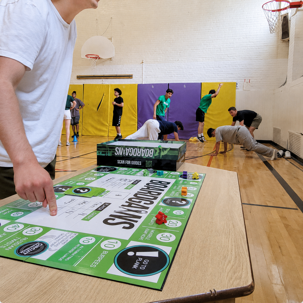 A person in a white shirt points at a green and white board game laid out on a table in a school gym, with several kids playing basketball and dodgeball in the background.