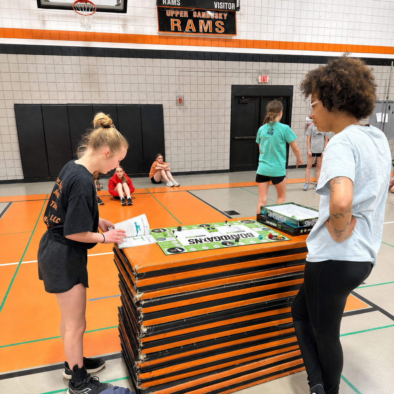 A stack of orange and black padded folding tables in a gymnasium, with two girls standing at the front and other girls in the background. A sign above reads 'RAMS' and 'VISITOR'.