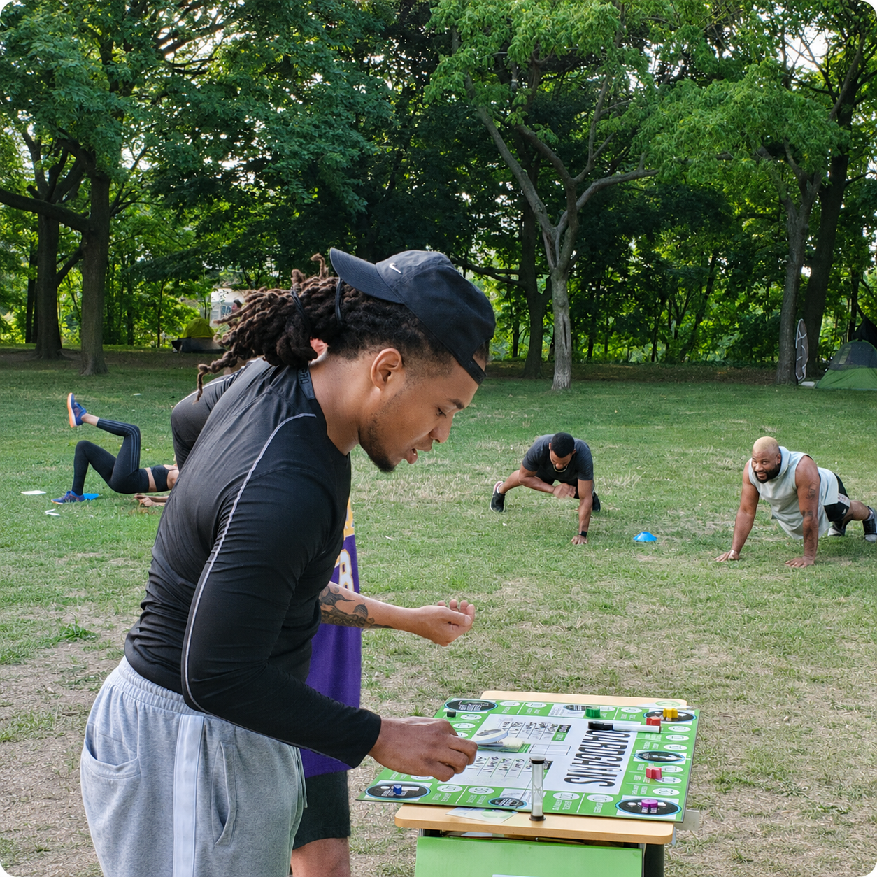 A man in a black long-sleeve shirt and gray sweatpants stands at a small table in a grassy park, playing a board game with a green and white game board. Other people are exercising in the background among trees.